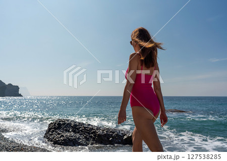 Beach Swimsuit Woman: Vacationer walks by sea's edge in pink swimwear under sunny skies in summer to relax. 127538285