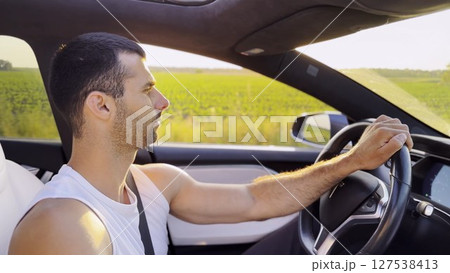 Profile of young handsome man driving a modern electric car with sunlight at background. Young male driver riding on electrical vehicle at countryside. Concept of journey and trip. Close up 127538413