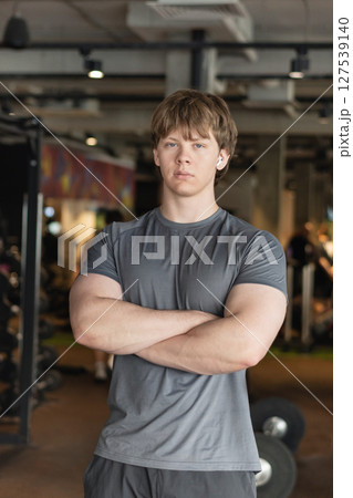 Sporty young personal trainer or fitness coach standing wearing gray t-shirt with cross hand at gym. Motivation for regular workout, body building, health and energy. Crafting my workout routine. Sporty young personal trainer or fitness coach standing wearing gray t-shirt with cross hand at gym. Motivation for regular workout, body building, health and energy. Crafting my workout routine. 127539140