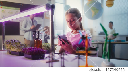 Talented Girl Sitting at Table and Using Smartphone, Conducting Biology Experiment on Plants 127540633