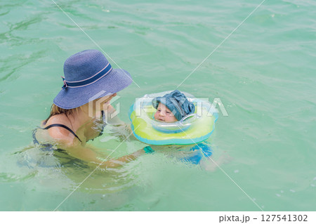 Baby floating in a neck ring in turquoise water with mom nearby. Early swimming and water safety concept Baby floating in a neck ring in turquoise water with mom nearby. Early swimming and water safety concept 127541302