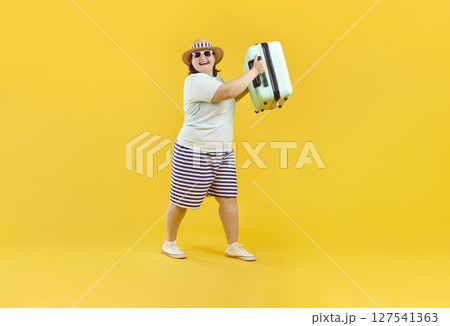 Fat funny woman in sunglasses carrying her suitcase on studio yellow background. 127541363