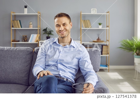 Confident attractive man sitting on sofa at home, looking at camera and smiling. 127541378