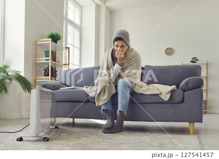 Young frozen man sitting wrapped in a blanket at home and trying to warm up with electric heater. 127541457