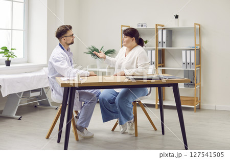 Young male doctor listening fat woman patient sitting in office during medical examination. Young male doctor listening fat woman patient sitting in office during medical examination. 127541505