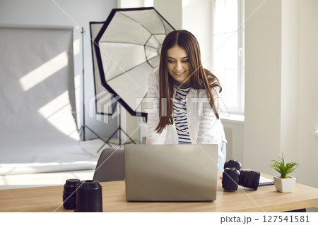 Young happy woman photographer standing at her workplace looking at the laptop and smiling. Young happy woman photographer standing at her workplace looking at the laptop and smiling. 127541581