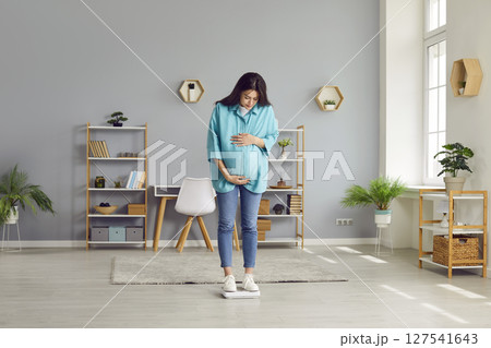 Portrait of a young pregnant woman measuring her weight standing on a scales at home. 127541643
