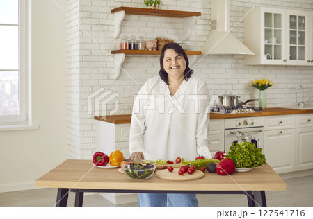 Cheerful overweight woman standing at table in kitchen cooking fresh vegetable salad Cheerful overweight woman standing at table in kitchen cooking fresh vegetable salad 127541716