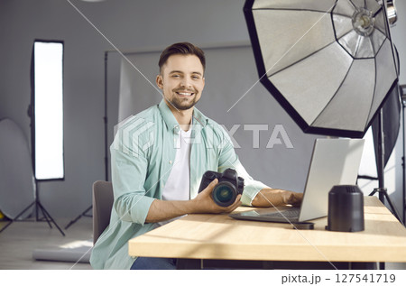 Young happy man photographer sitting at the desk with camera and lenses and smiling. Young happy man photographer sitting at the desk with camera and lenses and smiling. 127541719