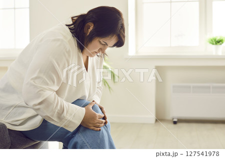 Young overweight woman holding a knee suffering from osteoarthritis sitting on sofa at home. 127541978