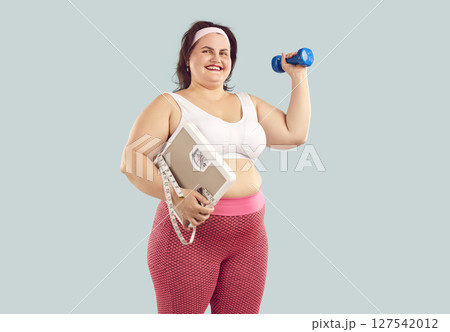 Happy fat woman wearing sportswear holding scales, dumbbell and measuring tape on blue background. Happy fat woman wearing sportswear holding scales, dumbbell and measuring tape on blue background. 127542012