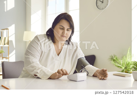 Overweight brunette woman measuring pressure while sitting at table at home 127542018