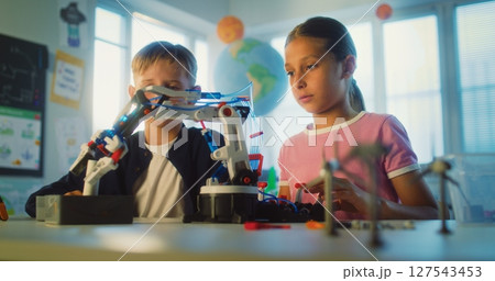 Team of Talented Boy and Girl Sitting at Table, Studying Robotic Arm Model During Science Lesson 127543453