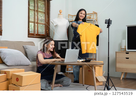 Asian woman friends couple talk to camera to sell the clothes at home. Young attractive business people female live streaming and showing clothing to customer in online Asian woman friends couple talk to camera to sell the clothes at home. Young attractive business people female live streaming and showing clothing to customer in online 127543519