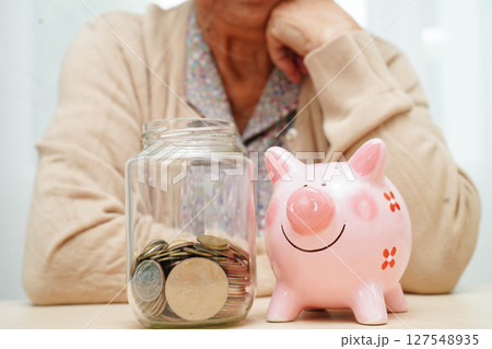 Retired elderly woman counting coins money with piggy bank and worry about monthly expenses and treatment fee payment. Retired elderly woman counting coins money with piggy bank and worry about monthly expenses and treatment fee payment. 127548935