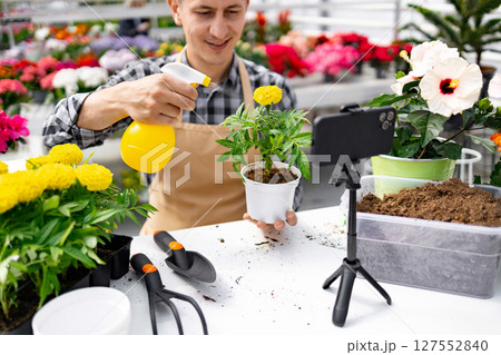 A florist sprays water on a plant, recording a gardening vlog using a smartphone on a tripod in a plant shop. 127552840