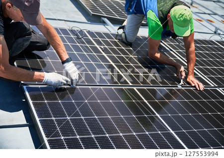 Workers building photovoltaic solar panel system on rooftop of house. Men engineers installing solar module with help of wrench outdoors. Alternative and renewable energy generation concept. 127553994