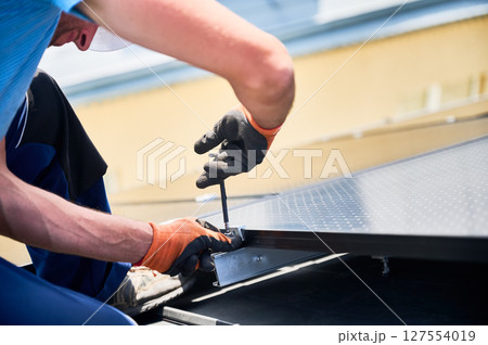 Worker building photovoltaic solar panel system on rooftop of house. Close up of man engineer in gloves installing solar module with help of hex key outdoors. Renewable energy generation concept. 127554019