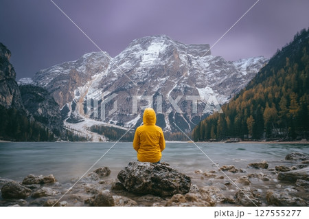 A man is seated on a rock at the edge of Braies Lake in Italy, with a backdrop of mountains and forest 127555277