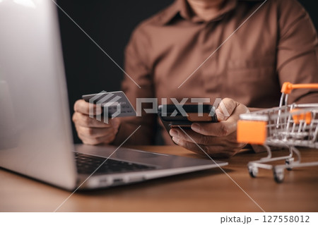 A man in a brown shirt makes an online payment using a credit card and smartphone while working on a laptop. A small shopping cart symbolizes digital shopping, e-commerce, and financial transactions. A man in a brown shirt makes an online payment using a credit card and smartphone while working on a laptop. A small shopping cart symbolizes digital shopping, e-commerce, and financial transactions. 127558012