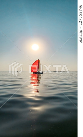 A sailboat with a vibrant red spinnaker sail glides smoothly across calm water under a clear sky. Silhouettes of people are visible on the boat, enjoying a peaceful sailing experience. The sun reflect 127558746