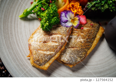 Pan-seared fish fillet with vibrant vegetables, including broccoli, cherry tomatoes, and carrot ribbons, garnished with edible flowers. Accompanied by a dark sauce jug. Fine dining closeup 127564152
