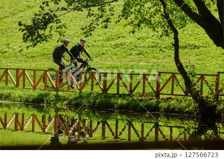 A blissful couple, adorned in professional cycling gear, enjoys a romantic bicycle ride through a park, surrounded by modern natural attractions, radiating love and happiness 127566723
