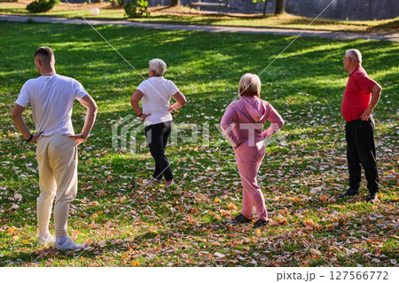 A group of seniors follows a trainer, engaging in outdoor exercises in the park, as they collectively strive to maintain vitality and well-being, embracing an active and health-conscious lifestyle in 127566772