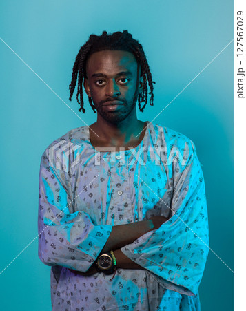 A Sudanese man adorned with modern dreadlocks stands proudly in traditional Sudanese attire, his arms crossed, conveying a blend of cultural heritage and contemporary style against a vibrant blue 127567029