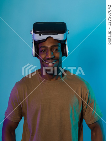 In a futuristic visual, an African American man stands isolated against a striking blue backdrop, adorned with VR glasses that transport him into a cutting-edge virtual reality experience, merging In a futuristic visual, an African American man stands isolated against a striking blue backdrop, adorned with VR glasses that transport him into a cutting-edge virtual reality experience, merging 127567042