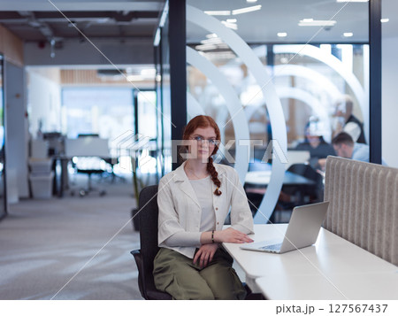 A young businesswoman with orange hair sitting confidently, fully engaged in her work on the laptop, exuding creativity, ambition, and a vibrant sense of individuality 127567437