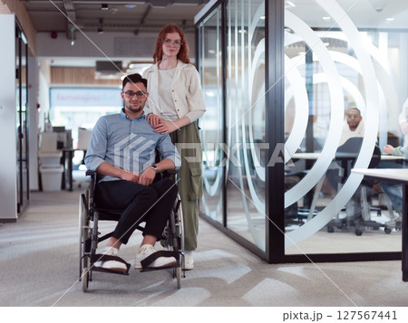 Young business colleagues, collaborative business colleagues, including a person in a wheelchair, walk past a modern glass office corridor, illustrating diversity, teamwork and empowerment in the 127567441