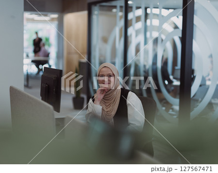 In a modern office, a young Muslim entrepreneur wearing a hijab sits confidently and diligently works on her computer, embodying determination, creativity, and empowerment in the business world 127567471