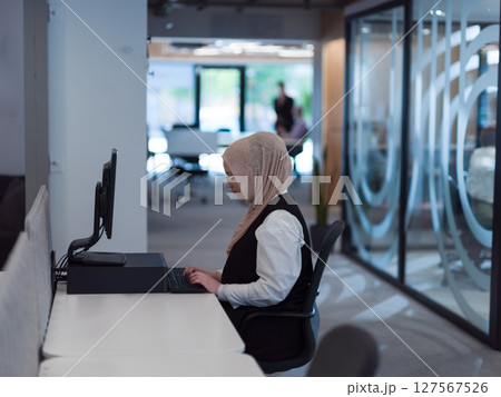 In a modern office, a young Muslim entrepreneur wearing a hijab sits confidently and diligently works on her computer, embodying determination, creativity, and empowerment in the business world In a modern office, a young Muslim entrepreneur wearing a hijab sits confidently and diligently works on her computer, embodying determination, creativity, and empowerment in the business world 127567526