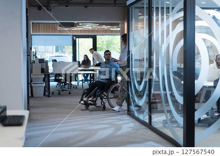 Young business colleagues, collaborative business colleagues, including a person in a wheelchair, walk past a modern glass office corridor, illustrating diversity, teamwork and empowerment in the 127567540