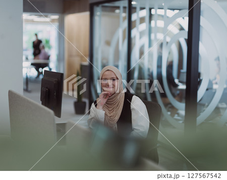 In a modern office, a young Muslim entrepreneur wearing a hijab sits confidently and diligently works on her computer, embodying determination, creativity, and empowerment in the business world 127567542