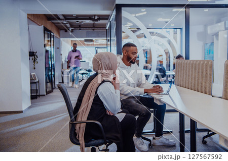 In a modern office setting, an African American businessman and his Muslim colleague, wearing a hijab, engage in collaborative discussions, tackling various business tasks and solving problems 127567592