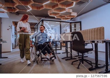 Young business colleagues, collaborative business colleagues, including a person in a wheelchair, walk past a modern glass office corridor, illustrating diversity, teamwork and empowerment in the Young business colleagues, collaborative business colleagues, including a person in a wheelchair, walk past a modern glass office corridor, illustrating diversity, teamwork and empowerment in the 127567596