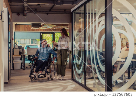 Young business colleagues, collaborative business colleagues, including a person in a wheelchair, walk past a modern glass office corridor, illustrating diversity, teamwork and empowerment in the Young business colleagues, collaborative business colleagues, including a person in a wheelchair, walk past a modern glass office corridor, illustrating diversity, teamwork and empowerment in the 127567605