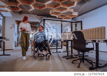 Young business colleagues, collaborative business colleagues, including a person in a wheelchair, walk past a modern glass office corridor, illustrating diversity, teamwork and empowerment in the Young business colleagues, collaborative business colleagues, including a person in a wheelchair, walk past a modern glass office corridor, illustrating diversity, teamwork and empowerment in the 127567628