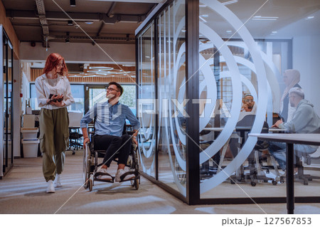 Young business colleagues, collaborative business colleagues, including a person in a wheelchair, walk past a modern glass office corridor, illustrating diversity, teamwork and empowerment in the 127567853