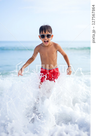Boy Enjoying Splashing Water at Beach on Sunny Day Wearing Sunglasses and Red Swim Trunks 127567864