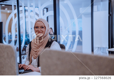 In a modern office, a young Muslim entrepreneur wearing a hijab sits confidently and diligently works on her computer, embodying determination, creativity, and empowerment in the business world 127568026