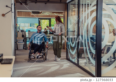 Young business colleagues, collaborative business colleagues, including a person in a wheelchair, walk past a modern glass office corridor, illustrating diversity, teamwork and empowerment in the 127568033