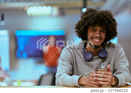In a modern startup office, an African American man, balancing work and technology, utilizes headphones and a smartphone, emblematic of contemporary multitasking and productivity 127568220