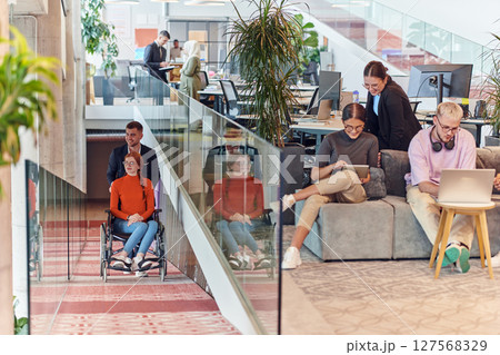 A company director assists his business colleague in a wheelchair, helping her navigate to their startup office, where they work alongside their diverse team of colleagues, emphasizing inclusivity and 127568329