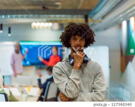 In a modern office environment, an African American young entrepreneur with headphones engages in work, while in the background, his dedicated colleagues exemplify teamwork and collaboration In a modern office environment, an African American young entrepreneur with headphones engages in work, while in the background, his dedicated colleagues exemplify teamwork and collaboration 127568340