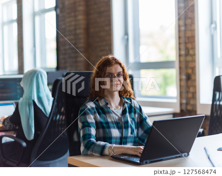 A young and successful businesswoman with vibrant orange hair engages in focused work within a modern office, showcasing her confidence and dynamic approach as she utilizes her laptop, emblematic of a 127568474