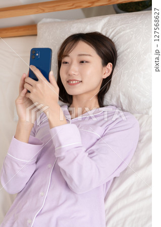 ベッドで寝たままスマホを操作する若い女性 ベッドで寝たままスマホを操作する若い女性 127568627