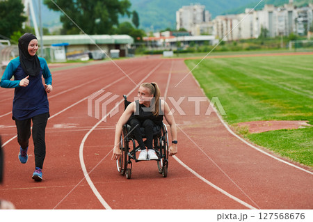 A Muslim woman in a burqa running together with a woman in a wheelchair on the marathon course, preparing for future competitions. 127568676